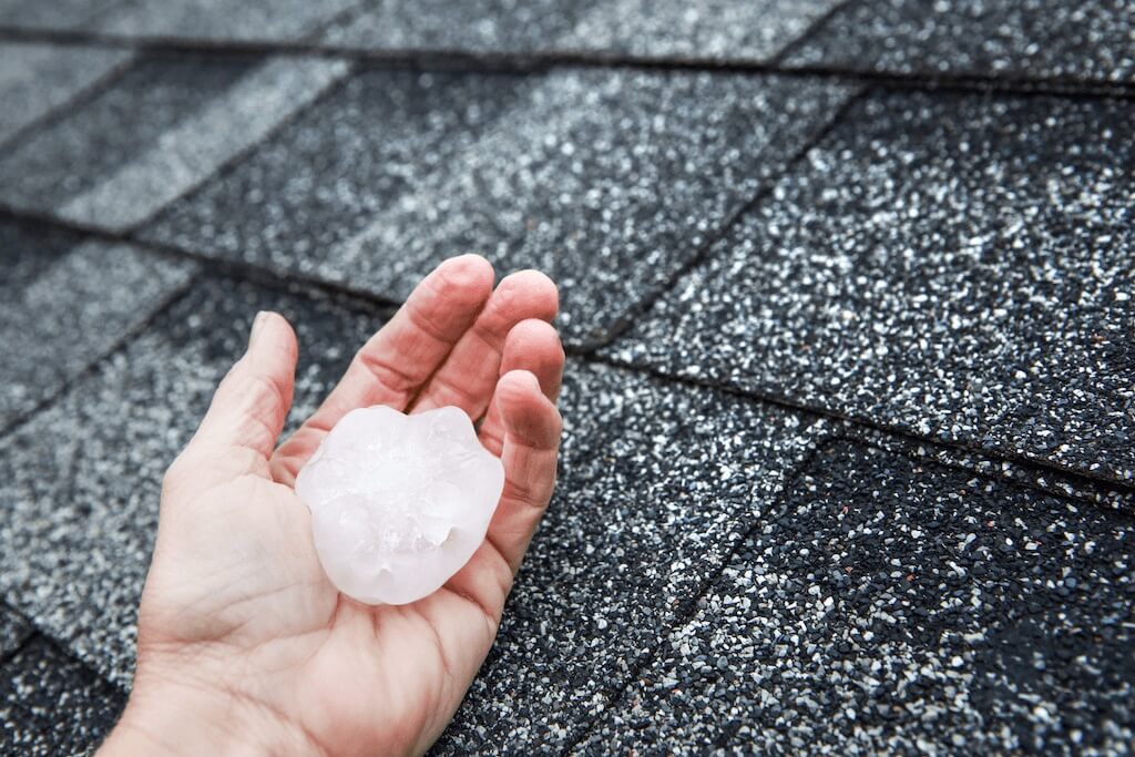 hand holding piece of hail in front of asphalt shingles on roof